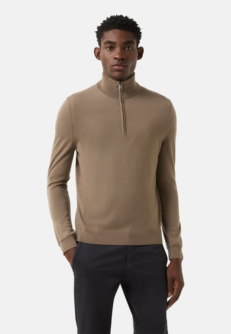 Brauner Reißverschluss-Pullover mit Rippenstrickmuster, hoher Kragen und langen Ärmeln, kombiniert mit dunkler Hose für Kontrast.