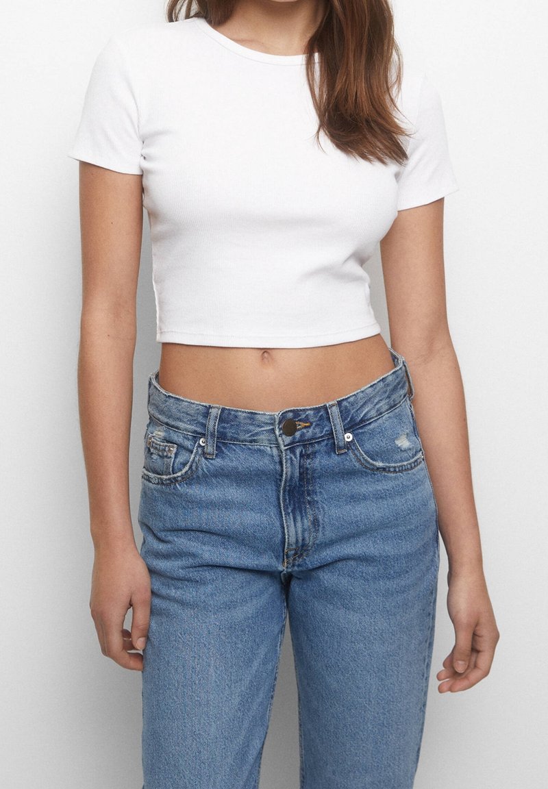 Femme portant un haut court blanc à manches courtes et un jean bleu taille haute, debout devant un fond clair uni.