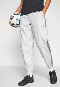 Šviesiai pilkos vėjo nepraleidžiančios kelnės su elastinėmis rankogaliais, šoniniais kišenėmis ir lygiu paviršiumi. Kairėje rankoje laikomas juodas futbolo kamuolys.