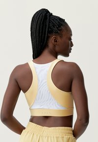 Sujetador deportivo con un suave frente amarillo, panel trasero de malla blanca, diseño de espalda tipo racerback y un sutil logotipo en la parte inferior. Material suave y elástico.