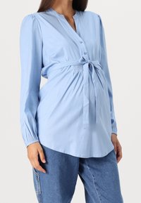 Světlemodrá těhotenská blůza s knoflíkovým zapínáním vpředu, dlouhými rukávy a látkovým opaskem v pase. Hladká textura, volný střih a zakřivený lem.