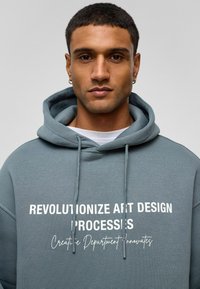 Hoodie in gedempt blauw-grijs met witte tekst: "REVOLUTIONIZE ART DESIGN PROCESSES" en "Creative Department Innovates." Zachte stof, relaxte pasvorm.