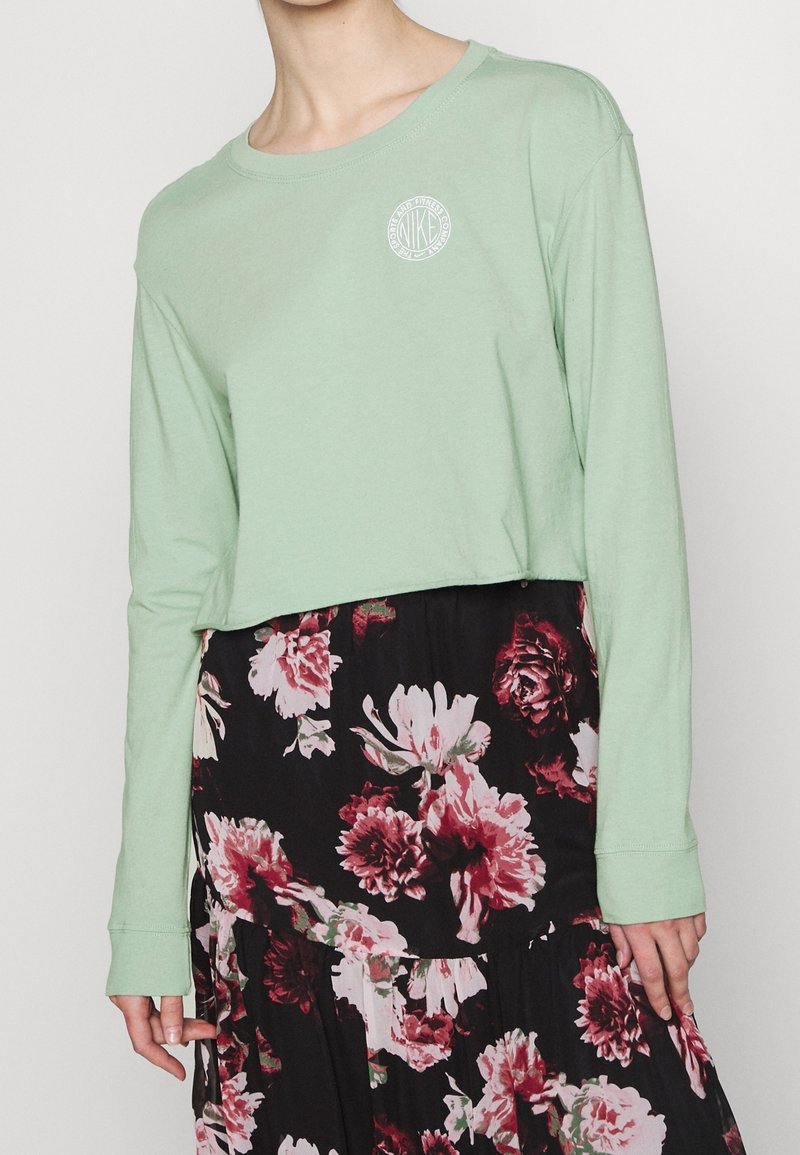 Světle zelená dlouhá rukávová košile z měkké bavlny s kulatým výstřihem a malým bílým logem na prsou, nošená přes květinovou sukni.