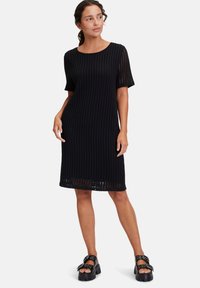 Schwarzes Strickkleid mit lockerer Passform, kurzen Ärmeln und einem strukturierten, offenen Muster. Getragen mit klobigen schwarzen Keilsandaletten.