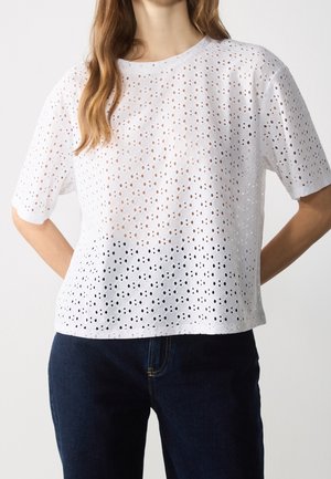 Vrouw draagt een witte blouse met korte mouwen van ooglet kant met cirkelvormige uitsnijdingspatronen, gecombineerd met donkerblauwe jeans.