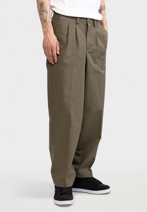 Mann iført olivengrønne plissert bukser, svarte sneakers og en hvit skjorte, stående med hendene i lommene mot en ensfarget bakgrunn.