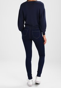 Marinblå pullover med puffade ärmar, kombinerad med tajta mörka jeans och vita sneakers med röda detaljer; slätt tyg och skräddarsydd passform.