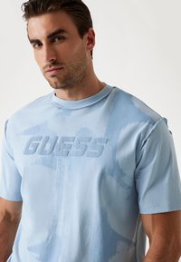 Tricou din bumbac de culoare albastru deschis, cu logo-texturat "GUESS", mâneci scurte, gât rotund și un model abstract în nuanțe variate de albastru.