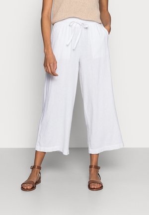 Weiße Leinen-Culottes mit lockerer Passform und verstellbarem Bund. Kombiniert mit braunen Sandalen mit Riemchen. Einfaches, unauffälliges Design.