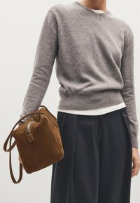 Pull en laine gris, t-shirt blanc, pantalon bleu marine et un sac en daim marron avec fermeture éclair et bandoulière réglable, tenu d'une main.