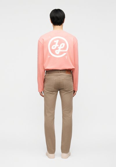 Άτομο με κοντά μαύρα μαλλιά που φοράει ροζ μακρυμάνικη μπλούζα με λευκό γραφικό κείμενο στην πλάτη και μπεζ παντελόνι ίσιας γραμμής.