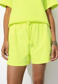 Shorts en coton jaune fluo avec une taille élastique et un cordon de serrage, comportant un petit patch en bas à droite. Texture douce.