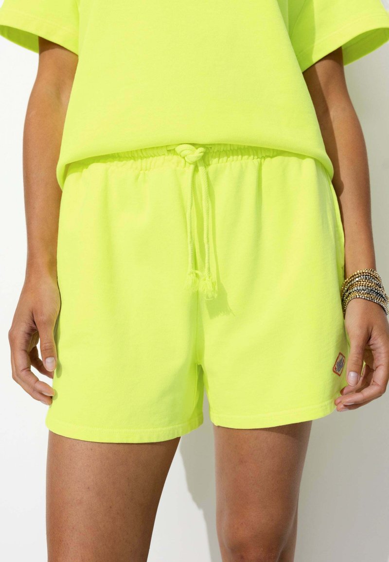Shorts en coton jaune fluo avec une taille élastique et un cordon de serrage, comportant un petit patch en bas à droite. Texture douce.