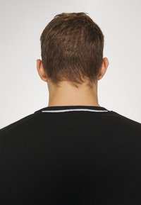 Homme aux cheveux bruns courts portant un pull noir avec une bande blanche au niveau de l'encolure, vu de dos sur un fond uni.