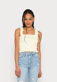 Top cropped amarelo com pregas, alças com babados, padrão texturizado e um design ajustado, combinado com jeans claros desgastados.
