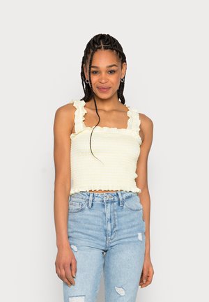 Top cropped amarelo com pregas, alças com babados, padrão texturizado e um design ajustado, combinado com jeans claros desgastados.