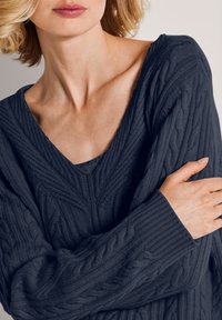 Frau mit heller Haut und blonden Haaren, die einen dunkelblauen, strukturierten Pullover trägt, die Arme vor der Brust verschränkt.
