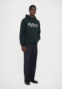 Dark green hoodie with white script logo, front pocket, and drawstring. Worn with pinstriped black pants and black shoes.