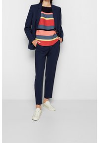 Vrouw in een marineblauwe pak, met een gestreepte multikleurtrui en witte sneakers, staand met handen in haar zakken tegen een witte achtergrond.