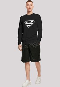 Black sweatshirt featuring a textured silver graphic of the Superman logo. Paired with loose black shorts and white sneakers.