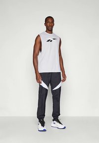 Vyras stovi vilkėdamas baltą be rankovių sportinį marškinėlį su juodu logotipu, juodas sportinėse kelnes su baltomis chevrono juostelėmis ir baltus sportinius batelius su mėlynais padais.