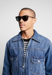 Jachetă din denim cu un spălare albastră, având un guler ascuțit, buzunare frontale și închidere cu fermoar. Ochelari de soare negri cu lentile întunecate.