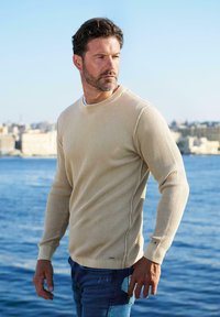 Beiger Ribbenpullover mit langen Ärmeln und weitem Rundhalsausschnitt, kombiniert mit blauen Jeans, vor einem Küstenhintergrund.