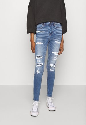 Vaqueros ajustados de color azul claro con rotos, combinados con zapatillas blancas y una camiseta negra de manga larga y holgada, de pie frente a un fondo sencillo.