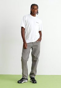 Hvit bomulls t-skjorte med logo, grå denim-bukser og svarte sneakers med hvite detaljer. Modellen står mot en ensfarget bakgrunn.
