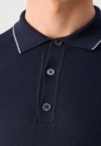 Polo en maille bleu marine avec trois boutons et un col présentant un liseré fin blanc sur le cou et le haut de la poitrine d'une personne.
