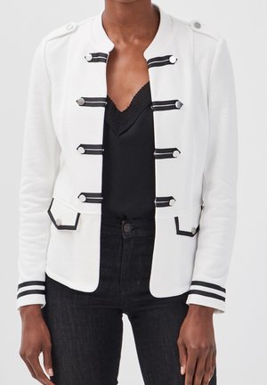 Femme portant une veste militaire blanche avec des boutons argentés et des finitions noires, associée à un caraco noir à dentelle et un pantalon noir.