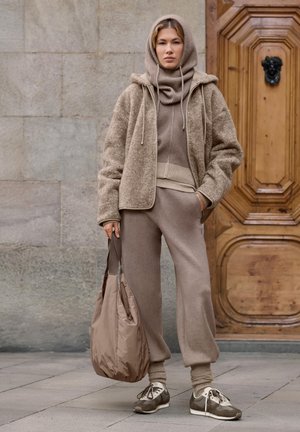 Femeie îmbrăcată în tricot bej stratificat și pantaloni joggers, ținând o geantă tote mare bej, stând în fața unei uși de lemn pe o pavaj de piatră.