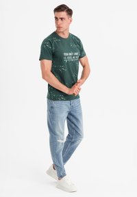 Camiseta de algodón verde con texto blanco, combinada con Jeans claros desgastados y zapatillas blancas, llevada por un hombre en un fondo neutro.