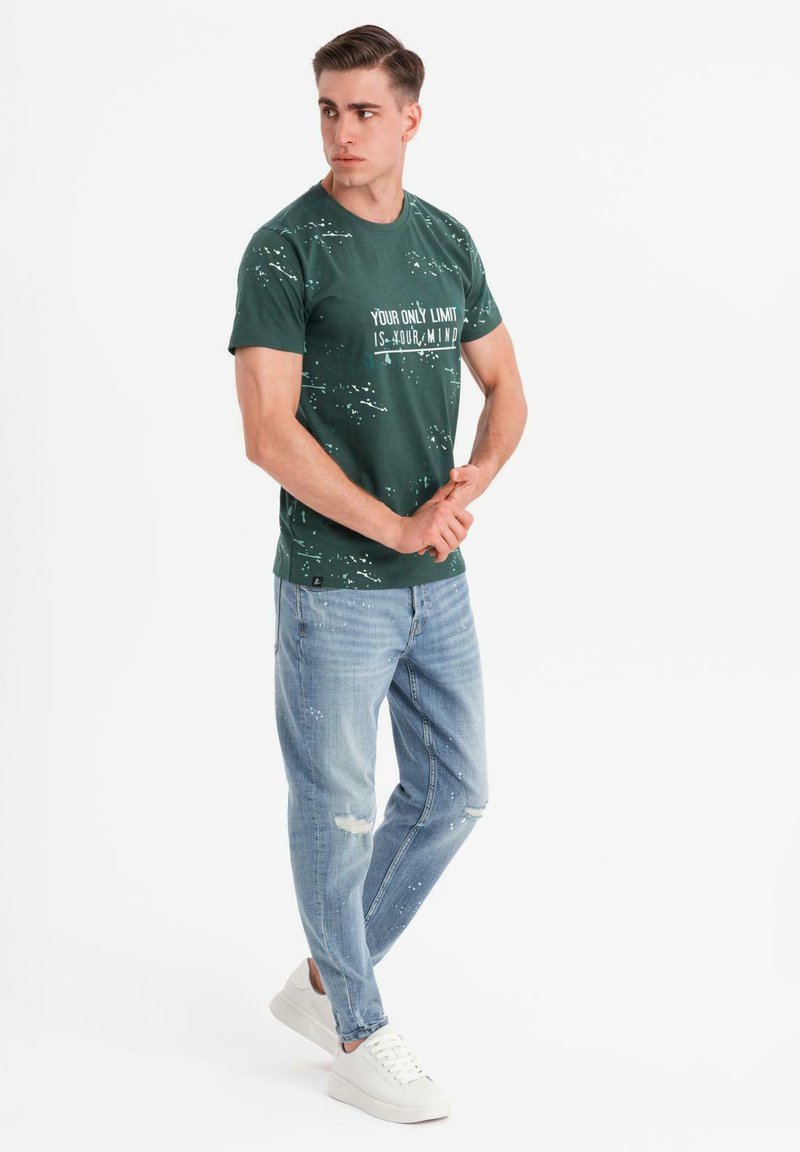 Camiseta de algodón verde con texto blanco, combinada con Jeans claros desgastados y zapatillas blancas, llevada por un hombre en un fondo neutro.