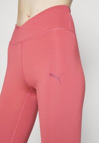 Lyserøde atletiske leggings med høj talje, lavet af glat stof, med flade sømme og et lille lilla Puma-logo på venstre lår.