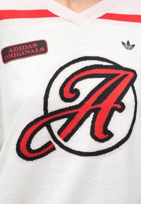 Pull blanc avec des rayures rouges, présentant un grand logo "A" noir et rouge brodé ainsi qu'un patch "ADIDAS ORIGINALS". Petit logo trèfle Adidas vert.