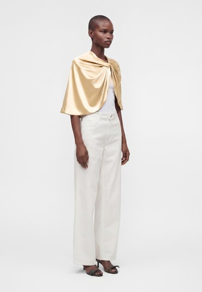 Femme portant une cape en satin doré brillant par-dessus un top blanc sans manches, un pantalon blanc à jambes larges et des talons ouverts noirs, debout devant un fond uni.