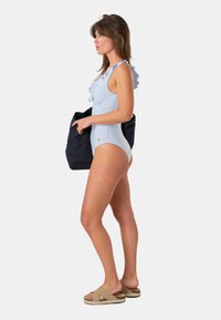 Femme en maillot de bain une pièce rayé bleu et blanc avec des volants, tenant un grand sac cabas noir, portant des sandales beige, faisant face à gauche.