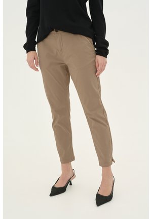 Personne portant un pantalon beige slim, un haut noir à manches longues et des escarpins noirs à talons avec bride arrière, debout sur un fond blanc.