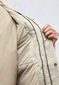 Beige isoleret jakke med en glat ydre tekstur og en skjult lynlås lomme. Indvendig har den kviltet foring og kontrast lynlås.