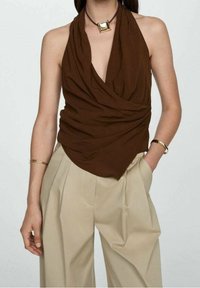 Top marrone senza maniche con tessuto drappeggiato e scollatura profonda, abbinato a pantaloni beige a vita alta. Accentuato da una collana con pendente quadrato in oro.