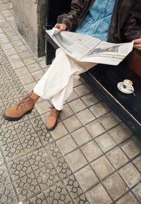 Brązowe zamszowe buty na sznurówki z grubą podeszwą, zestawione z szerokimi, kremowymi spodniami. Gazeta jest trzymana nad kubkiem kawy na kafelkowanej powierzchni.