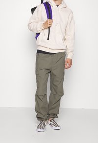 Sudadera beige con un bolsillo frontal, pantalones de cargo oliva con bolsillos laterales, y zapatillas de cuadro negro y blanco sin cordones. Mochila con correas moradas.