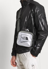 Giacca nera lucida con dettagli in rete, abbinata a una borsa rettangolare argentata con la scritta "THE NORTH FACE extreme" in lettere nere.