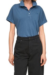 Modré polo tričko s krátkým rukávem a špičatým límcem, s trojicí knoflíků a malým logem na hrudi, v kombinaci s černými kalhotami.