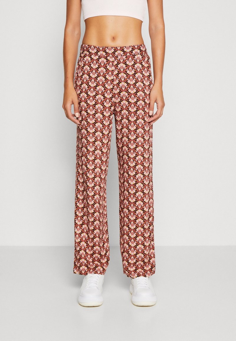 Femme portant un pantalon taille haute à jambes larges avec un motif floral géométrique rouge et rose et des baskets blanches, debout contre un mur uni.