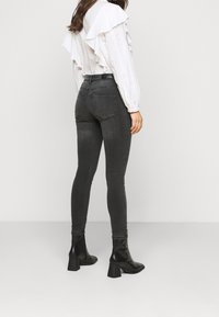 Dunkelgraue Skinny-Jeans mit hoher Taille und glatter Textur, kombiniert mit einer weißen Rüschenbluse und schwarzen Ankle-Boots mit Absatz.