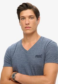 Maglietta blu a V in tessuto morbido con maniche corte e un piccolo logo nero sul petto. Modello con le braccia incrociate. Orologio argento al polso.