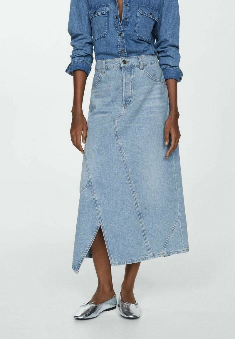 Femme portant une jupe midi en denim clair avec une fente à l'avant, une chemise en denim boutonnée et des ballerines argentées métalliques, debout devant un fond blanc.