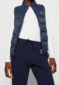 Granatowa kamizelka ocieplana z wysokim kołnierzem i zamkiem błyskawicznym, noszona na białej koszuli z długim rękawem, zestawiona z ciemnoniebieskimi spodniami dresowymi.